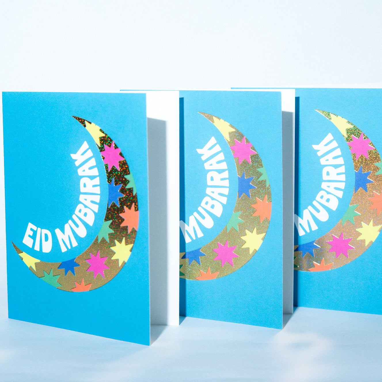 Three blue greeting cards with colorful crescent moon designs and 'Eid Mubarak' text on a light blue background.