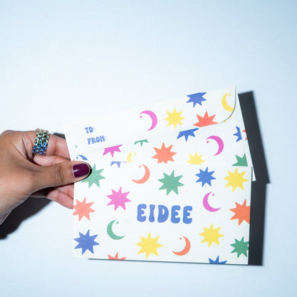 Hand holding a colorful card with star and moon patterns and the name 'EIDEE' on a light blue background.