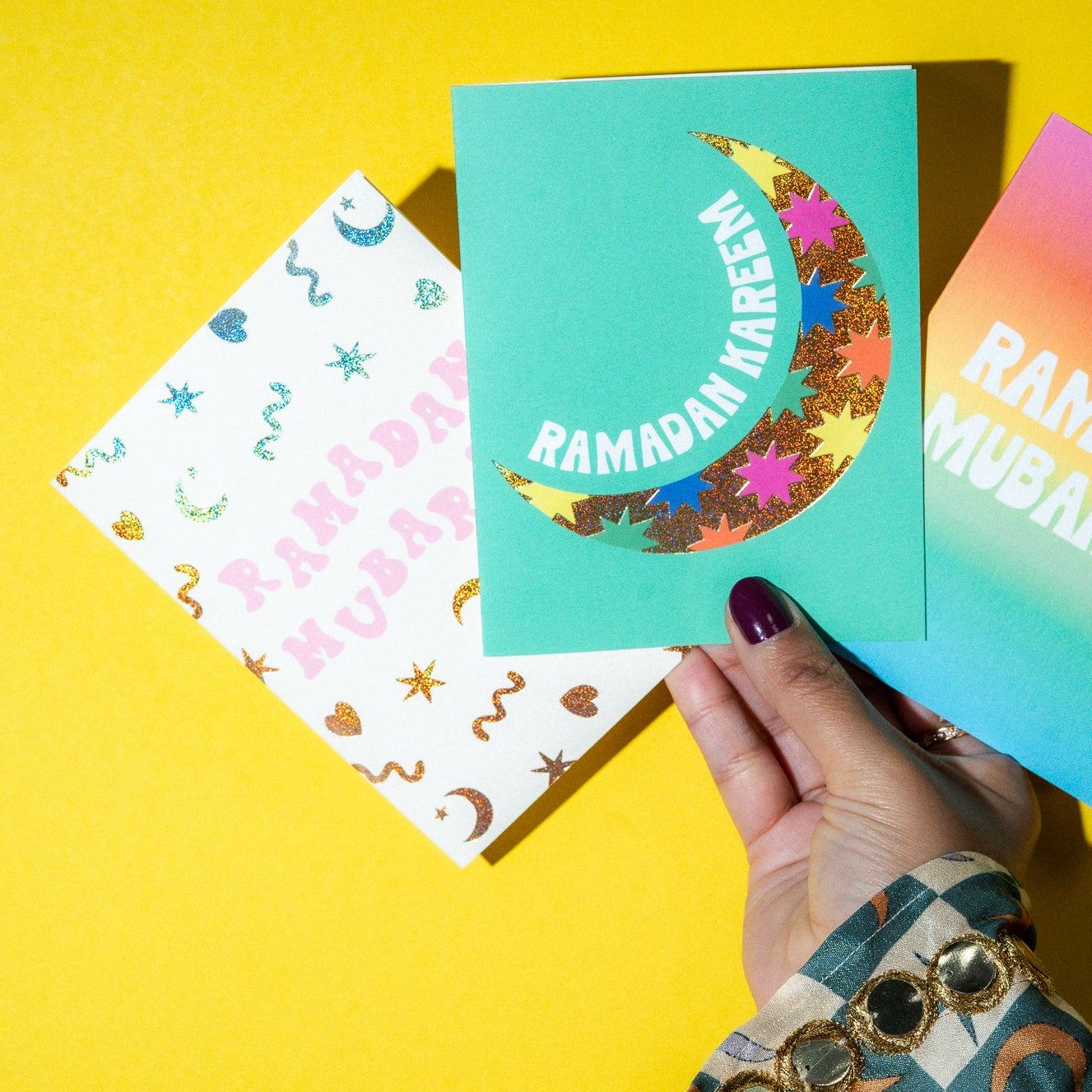 Three colorful Ramadan greeting cards held by a hand on a yellow background